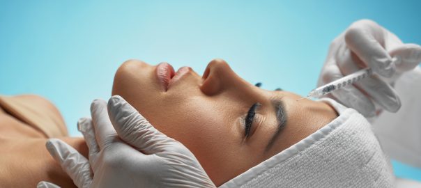 Close up of botox injection in female forehead. Side view of cosmetologist using syringe with special liquid, holding chin while patient with towel on head lying. Concept of cosmetology, beauty.